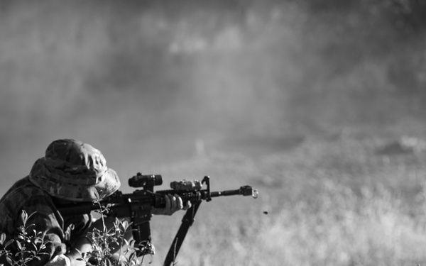 A black and white photograph showing a person in camouflage military attire and a boonie hat in a prone or crouched firing position. The individual is aiming a rifle equipped with an optical scope and bipod across an open field. The shooter is partially concealed by low brush and vegetation in the foreground.