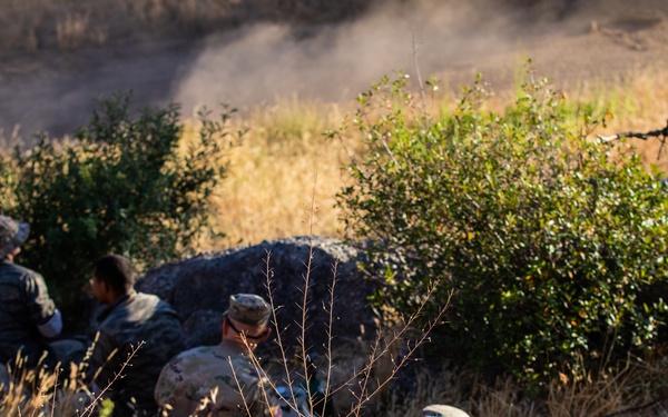 The image shows several individuals in military-style camouflage uniforms positioned behind rocks and sparse vegetation in an arid, dry grassland environment. They appear to be taking cover or observing from a concealed position. The terrain is characterized by dry golden grass, shrubs, and rocky outcroppings.