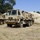 The image shows military medical vehicles parked on dry grassland, identifiable by the prominent Red Cross markings on their cargo areas. The vehicles appear to be FMTV (Family of Medium Tactical Vehicles) or similar heavy tactical trucks in desert tan/sand camouflage paint. A large inflatable tan-colored structure, likely a field hospital or medical tent, is visible being deployed or stored behind the lead vehicle.
