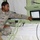 A U.S. Army soldier identified by name tape as 'SCOTT' is operating or demonstrating a military field equipment console inside what appears to be a tent or temporary shelter. The soldier is wearing Army Combat Uniform (ACU) with a Sergeant rank patch and U.S. flag patch on the left shoulder. He is pointing at a touchscreen display mounted on a light green military equipment rack.