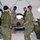 Military medical personnel are conducting what appears to be a field medical training exercise inside a tactical tent. Four uniformed soldiers — including Canadian Armed Forces members (identifiable by Canadian flag patches) and at least one US Army soldier — are attending to a patient lying face-down on a field stretcher or medical table. The setting is a portable military medical facility equipped with lighting, medical equipment, and field gear.