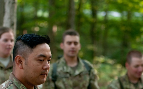 A group of U.S. Army soldiers in MultiCam (OCP) camouflage uniforms are gathered outdoors in a wooded area. The foreground soldier, whose name tape reads 'ESEBAGIRA' (or similar), wears a U.S. flag patch and a 3rd Infantry Division patch on the left sleeve. The soldiers appear to be at attention or in formation during a field exercise or briefing.