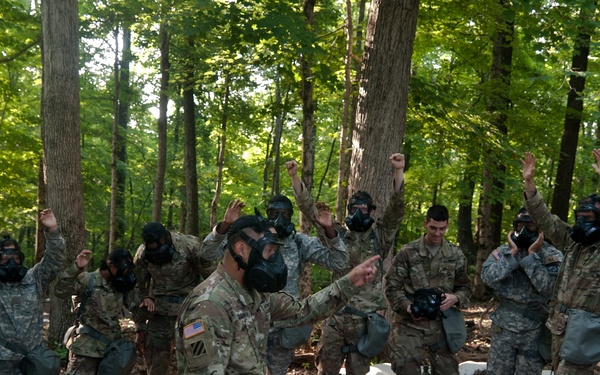 A group of military personnel in camouflage uniforms are participating in what appears to be a CBRN (Chemical, Biological, Radiological, Nuclear) training exercise in a wooded outdoor setting. Most soldiers are wearing M40-series gas masks, with several raising their hands or fists, while a central instructor figure points and directs the group. Equipment including gas mask carriers and protective gear bags are scattered on the ground around them.