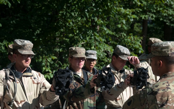 A group of military personnel in mixed camouflage uniforms are gathered outdoors in a wooded area. An instructor or senior soldier in OCP (Operational Camouflage Pattern) uniform appears to be conducting an equipment inspection or demonstration, handling what appears to be a gas mask or similar protective gear. The trainees are wearing various older-pattern uniforms including Desert Camouflage Uniform (DCU) and Woodland BDU patterns, suggesting this may be a training exercise.