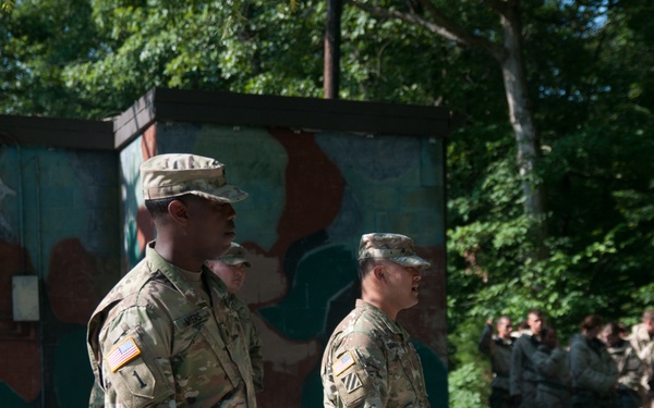 The image shows U.S. Army soldiers in MultiCam (OCP) camouflage uniforms standing at what appears to be a military training area or installation. Two soldiers are prominently visible in the foreground, with a larger group of soldiers assembled in the background. The setting includes a gravel surface, a painted structure, and a wooded area.