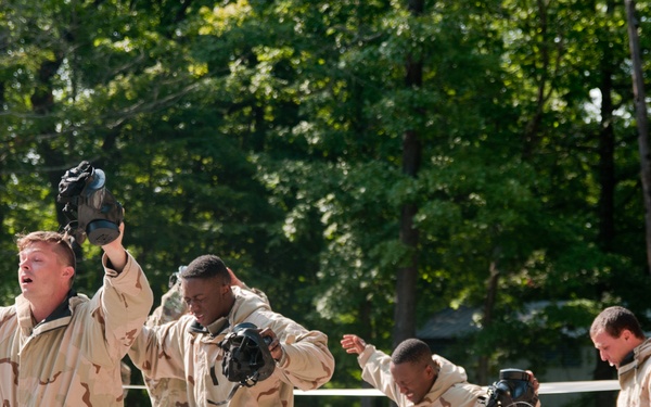 Four military personnel in desert camouflage uniforms are exiting what appears to be a gas chamber training exercise, holding their gas masks away from their faces. The individuals display visible physical reactions consistent with CS gas or similar irritant exposure, including raised arms and distressed expressions. The setting is an outdoor military training facility surrounded by trees.