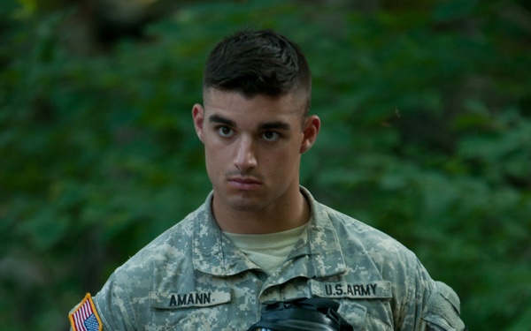 A U.S. Army soldier named Amann is photographed in Army Combat Uniform (ACU) digital camouflage pattern, holding a military gas mask (M40 or M50 series). He is standing in front of a green, leafy background and looking directly at the camera with a serious expression. The image appears to be a professional or semi-professional military portrait.