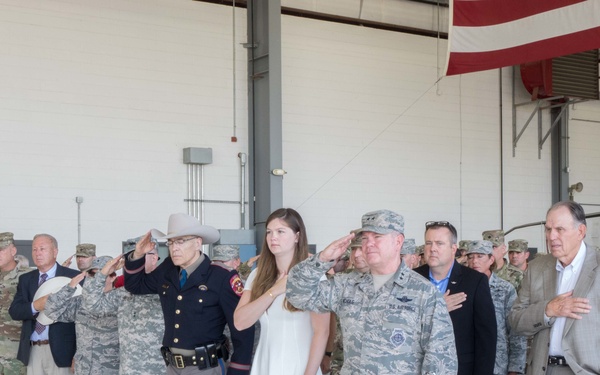 A formal military ceremony is taking place inside what appears to be an aircraft hangar. A mixed group of military personnel in camouflage uniforms, a law enforcement officer in a tan cowboy hat, and civilians are standing at attention — some saluting and some with hands over hearts — likely during the playing of the national anthem or Pledge of Allegiance. A partial American flag is visible in the upper right corner of the image.