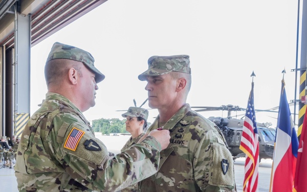 A U.S. Army military ceremony taking place inside an aircraft hangar, where one soldier is pinning a medal or award onto another soldier's uniform. Both individuals are wearing Army Combat Uniforms (ACU) in MultiCam/OCP pattern. The setting includes flags, a military helicopter, and what appears to be a military band in the background.