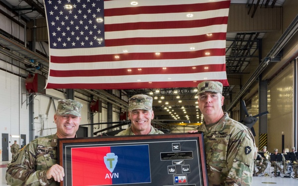 Three U.S. Army soldiers in OCP (Operational Camouflage Pattern) uniforms pose together holding a framed commemorative shadow box display inside a military hangar. A large American flag is draped prominently in the background. The setting appears to be a formal military ceremony, likely a change of command or unit recognition event.