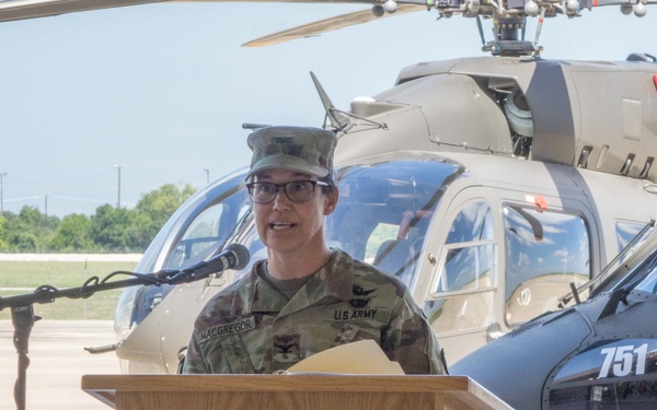 A U.S. Army officer identified by name tape as 'MACGREGOR' stands at a wooden podium bearing the seal of the State of Texas, delivering remarks at what appears to be an official military ceremony. The officer is wearing Operational Camouflage Pattern (OCP) uniform with colonel rank insignia (eagle) and aviator wings. A military helicopter numbered '751' is visible on the tarmac in the background.