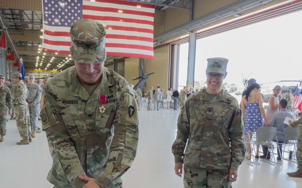A military ceremony taking place inside an aircraft hangar where two Army soldiers in OCP (Operational Camouflage Pattern) uniforms are cutting cakes with a saber. The cakes are decorated with unit insignia and read 'COL MACGREGOR' and 'COL BURKETT' along with 'CONGRATULATIONS,' suggesting a change of command or retirement ceremony. A large American flag is displayed in the background, and numerous other soldiers and civilians are present in the hangar.