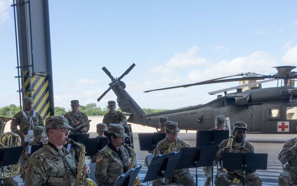 A United States Army military band performs inside or at the entrance of a hangar, with soldiers in camouflage uniforms playing brass and woodwind instruments from sheet music on stands. In the background, a UH-60 Black Hawk helicopter marked with a Red Cross medical insignia and 'United States Army' text is visible on the tarmac. The setting appears to be a formal or ceremonial musical performance at a military airfield.