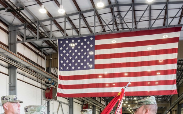 A U.S. Army military ceremony is taking place inside a large hangar with a prominent American flag displayed on the back wall. A soldier in the center is holding a furled unit guidon/colors, appearing to be in the process of a change of command or activation ceremony. Multiple soldiers in OCP (Operational Camouflage Pattern) uniforms are participating, with a formation of troops visible in the background.