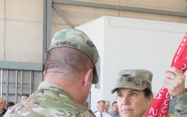 Two U.S. Army soldiers in OCP (Operational Camouflage Pattern) uniforms are participating in what appears to be a military colors/guidon ceremony, likely a change of command or unit activation/inactivation. The soldier facing the camera is handling a red guidon/streamer with white text visible, while the soldier with the name tape 'HENRY' has their back to the camera. Civilian observers are visible in the background.