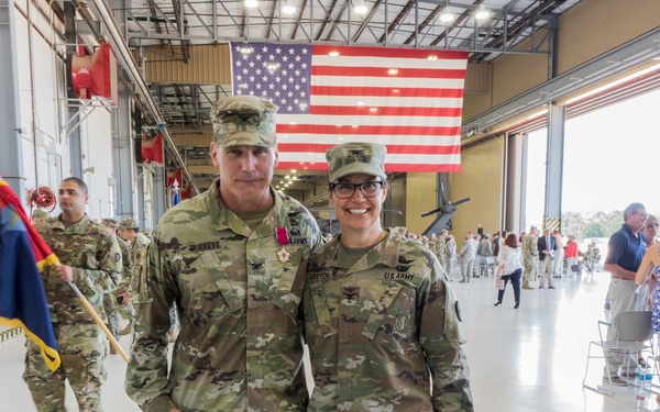 Two U.S. Army soldiers in OCP (Operational Camouflage Pattern) uniforms pose together in front of a large American flag inside a military hangar. A 'Farewell' cake and a ceremonial saber are visible in the foreground, indicating this is a farewell or change-of-command ceremony. The background shows additional soldiers, civilians, and what appears to be a helicopter in the hangar.