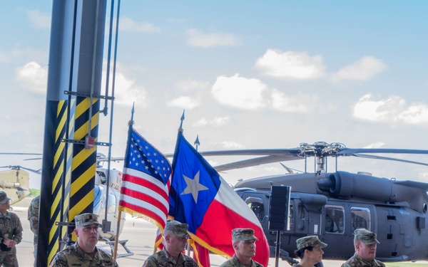 A military ceremony is taking place inside or at the entrance of an aviation hangar, with five uniformed soldiers seated in a row on folding chairs. The American flag and Texas state flag are displayed prominently behind the seated personnel, indicating a formal military event likely associated with the Texas National Guard or a Texas-based Army unit. Multiple military helicopters, including what appear to be UH-60 Black Hawks and CH-47 Chinooks, are visible on the flight line in the background.