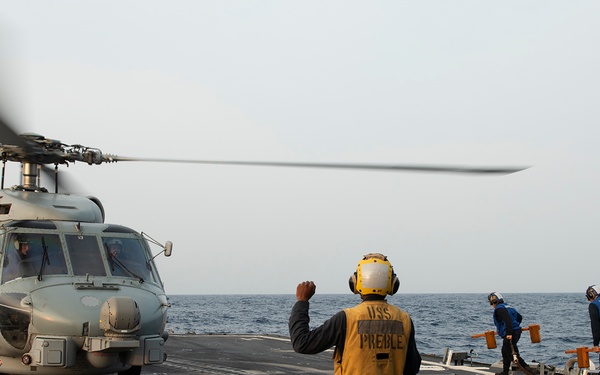 A naval flight deck crew member wearing a yellow vest marked 'USS PREBLE' is directing a military helicopter on the flight deck of a U.S. Navy destroyer. The helicopter, appearing to be an MH-60 Seahawk variant, has its rotors spinning and is positioned on the marked landing pad. Two additional crew members are visible in the background near the ship's stern railing.