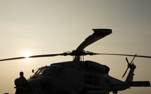 A military helicopter is parked on the flight deck of a naval vessel at sunset or sunrise, with a single person standing beside it in silhouette. The scene is captured in strong backlit conditions, creating dramatic silhouettes against a warm, hazy sky over open water. The helicopter appears to be a twin-engine naval variant, consistent with a Sikorsky SH-60/MH-60 Sea Hawk type.