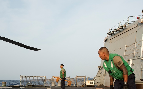 Two naval personnel wearing green safety vests are working on the flight deck of a U.S. Navy warship at sea. They appear to be handling helicopter tie-down chocks and chains, likely preparing or securing a helicopter on the deck. A helicopter rotor blade is partially visible in the upper left corner of the frame.