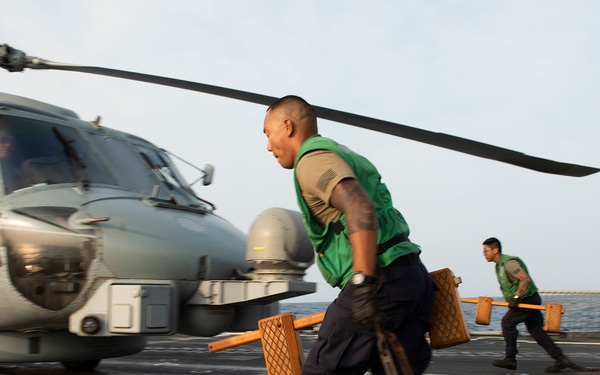 Two naval deck crew members wearing green safety vests are running across a ship's flight deck carrying wheel chocks away from a military helicopter marked '40'. The helicopter, identifiable as a Sikorsky MH-60R Seahawk, has its rotors spinning and a pilot visible in the cockpit. The scene depicts a standard flight deck operation, likely a helicopter launch or recovery evolution aboard a U.S. Navy vessel.