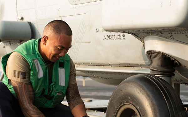 A military aviation technician wearing a green safety vest is crouched down working on the landing gear or tie-down chain of a military aircraft on what appears to be a naval vessel flight deck. The individual is using a tool to secure or release a chain attached to the aircraft's wheel assembly. The setting is consistent with carrier deck operations, with visible deck markings and aircraft components in the background.