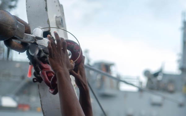 A worker wearing a blue hard hat and blue coveralls is reaching upward with both arms to handle rigging or chain equipment on what appears to be a naval vessel or port facility. The background shows the gray hull and superstructure of a large ship, suggesting a dockside or shipboard maintenance operation. The worker appears to be securing or adjusting a shackle, hook, or chain connection overhead.