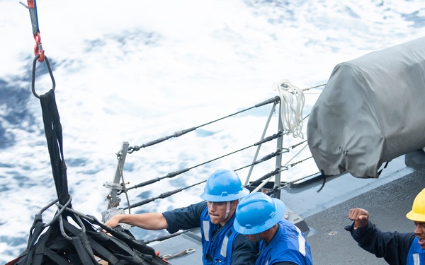 Naval personnel are conducting an underway replenishment (UNREP) operation on the deck of a U.S. Navy vessel at sea. Sailors wearing blue coveralls with reflective vests and hard hats are handling a cargo net loaded with boxed supplies being transferred via crane or highline. The rough sea conditions and ship railing are visible in the background.