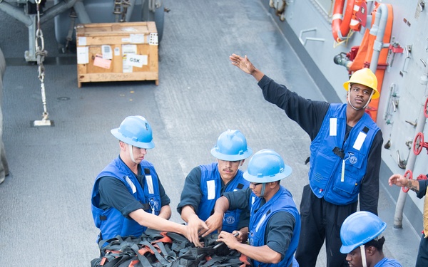 A group of U.S. Navy sailors aboard USS Preble are conducting a replenishment-at-sea (RAS) or cargo handling operation on the ship's deck. The sailors are working together to manage a large pallet of wooden crates secured with black cargo netting and orange/red straps. One sailor wearing a yellow hard hat appears to be directing the operation while others in blue vests and blue hard hats handle the cargo.