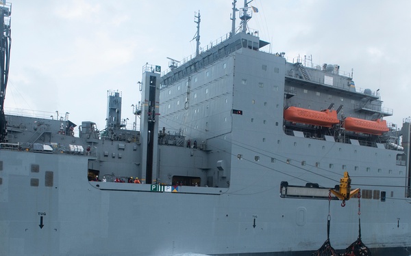 A large gray naval auxiliary or replenishment vessel is underway at sea in rough conditions, with white-capped waves visible alongside the hull. A cargo net loaded with supplies is suspended from a crane or highline transfer rig on the ship's side, indicating an underway replenishment (UNREP) operation in progress. The overcast sky and churning seas suggest challenging weather conditions during this logistics transfer.