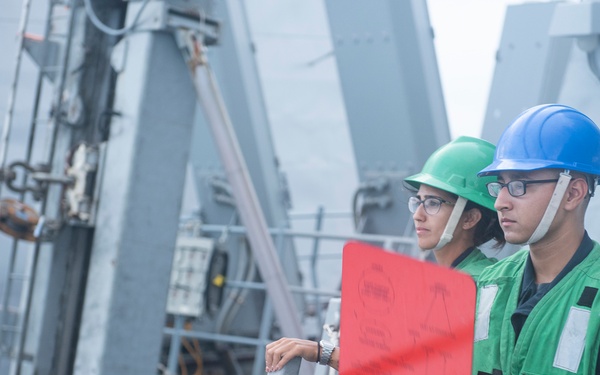 Two naval personnel wearing green safety vests and hard hats (one green, one blue) are standing on the deck of a military vessel. The individual in the foreground is holding a red signaling board, suggesting they are engaged in a replenishment-at-sea (RAS) or ship-to-ship communication operation. A third person wearing a white hard hat is partially visible in the background.