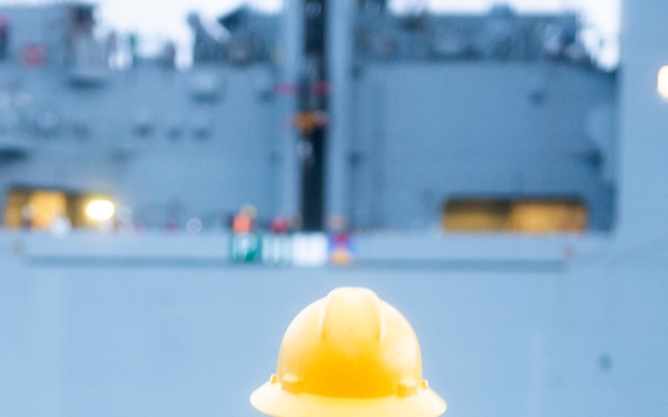 A sailor or crew member is seen from behind, wearing a yellow hard hat and an orange safety vest stenciled with 'USS PREBLE' and the number '35'. The individual is standing on a deck facing a large gray naval vessel in the background. The scene appears to be a maritime operation, likely a replenishment-at-sea or docking maneuver during low-light conditions.