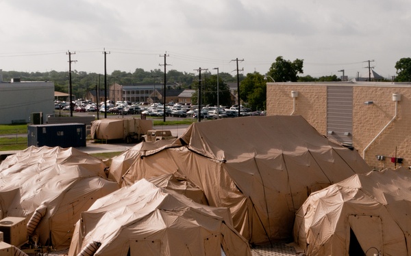An elevated view of a military tactical operations complex consisting of multiple interconnected tan/desert-colored inflatable or frame-supported tents deployed in a parking lot or hardstand area adjacent to permanent buildings. The setup includes satellite communication dishes, HVAC/climate control units, power generation equipment, and military vehicles, suggesting a command and control or communications node. The installation appears to be on a U.S. military installation based on the surrounding infrastructure.