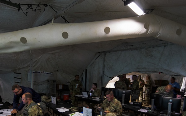 Military personnel are working inside a large inflatable command post tent equipped with computers, laptops, and communications equipment. Soldiers in camouflage uniforms are seated at long black tables operating workstations, suggesting a tactical operations center (TOC) or command and control exercise. The environment appears to be a field exercise or training event based on the temporary tent structure and equipment layout.