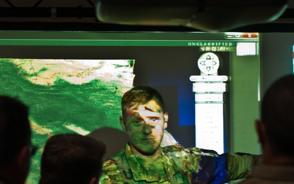 Military personnel are gathered in what appears to be a command operations center or briefing room. A soldier standing at the front is conducting a briefing, with projected map imagery and data displays visible on large screens behind him. The projection overlaps onto the briefer's body, creating a visual effect where the map imagery is cast across his face and uniform.