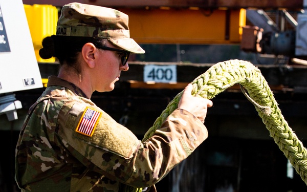 A female U.S. Army soldier in OCP (Operational Camouflage Pattern) uniform is handling a thick braided green mooring rope, likely during a watercraft or maritime operation. She is wearing sunglasses and a patrol cap, with a U.S. flag patch visible on her right shoulder. A second individual in a white t-shirt is partially visible to the right, and industrial port or dock equipment is visible in the background.