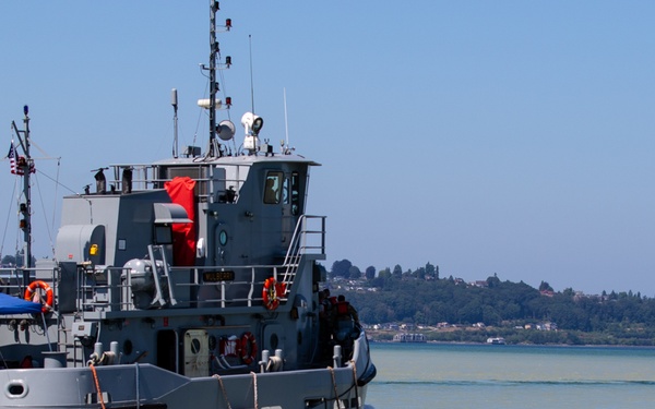 A U.S. Army vessel designated ST-914 is underway on a body of water, likely a bay or river estuary based on the greenish-brown water color. The gray military tugboat is accompanied by a smaller vessel with a blue canopy tent, with personnel visible on both crafts. A treeline and residential area are visible on the distant shoreline under clear blue skies.