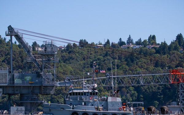 The image shows a military or industrial waterfront scene featuring a gray vessel or work boat moored alongside a large fixed crane structure with an extended boom painted in red and white. The setting appears to be a harbor or bay with a tree-covered hillside and residential structures visible in the background. The scene suggests an active naval or port facility with heavy lifting or loading equipment.
