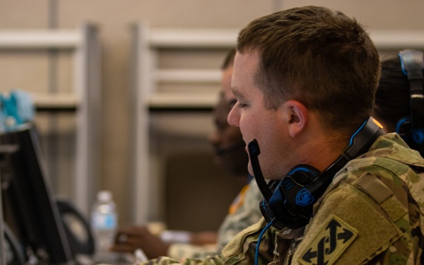 A U.S. Army soldier in OCP (Operational Camouflage Pattern) uniform is working at a computer workstation, wearing a headset with a boom microphone, suggesting a command post or operations center environment. The soldier has a unit patch visible on the left shoulder, identifiable as the 2nd Infantry Division patch. Multiple personnel and workstations are visible in the background, indicating a collaborative military operations or training environment.