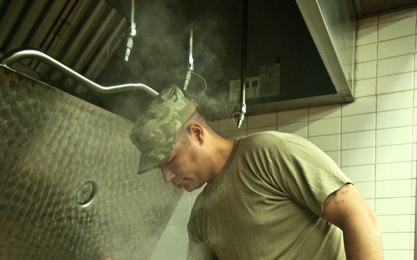 A U.S. Army soldier in OCP (Operational Camouflage Pattern) uniform and patrol cap is working in a military dining facility kitchen. He is leaning over a large commercial steam kettle, using a wire whisk to stir its contents. Steam is visibly rising from the cooking vessel, indicating active food preparation.