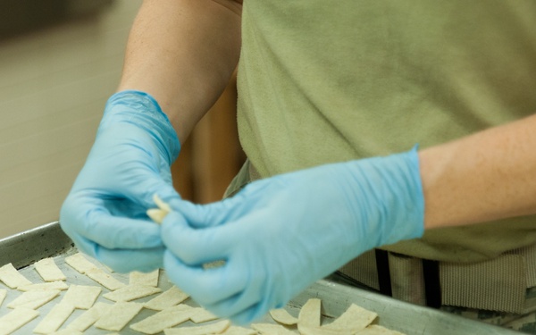 A person wearing blue latex gloves and military-style camouflage pants is arranging or sorting small flat pieces of dough or food items on a large metal baking sheet. The setting appears to be a commercial or institutional kitchen. The activity suggests food preparation, likely cutting or laying out dough pieces prior to baking.