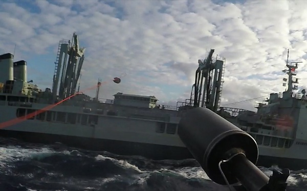 A naval replenishment-at-sea (RAS) operation is underway between two military vessels in rough open ocean conditions. A large auxiliary/supply ship is visible on the left, connected to a smaller warship on the right via a transfer line, with a red hose or cable visibly stretched between them. The image is captured from a low deck-level perspective, likely from a third vessel or the receiving ship, with a probe or nozzle apparatus in the foreground.