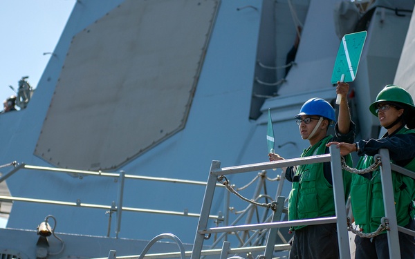 Two naval personnel wearing green safety vests and hard hats are standing on an elevated platform/gangway of a military vessel. One individual wearing a blue hard hat and one wearing a green hard hat appear to be conducting signaling or communication duties, with one holding up what appears to be a green paddle or signaling board. The setting is the exterior deck of a gray naval warship under clear blue skies.
