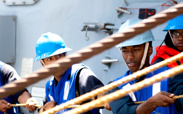 Four workers wearing blue hard hats and blue safety vests are pulling thick rope lines on what appears to be the deck of a naval or commercial vessel. Three individuals are actively hauling lines while a fourth, wearing a red life vest over their blue vest, observes or assists from behind. The scene depicts a mooring or line-handling operation typical of maritime deck work.
