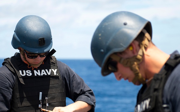 Two U.S. Navy sailors are on the deck of a naval vessel at sea, handling a belt of ammunition. Both personnel are wearing blue helmets, sunglasses, black tactical vests over Navy-issued shirts, and are engaged in what appears to be weapons preparation or maintenance. The ocean and partly cloudy sky are visible in the background.