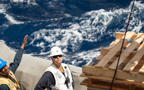 Four personnel are gathered on the deck of a vessel at sea, engaged in what appears to be a cargo or replenishment-at-sea operation. A stacked pile of wooden pallets secured by rope is visible to the right, while rough ocean water is visible in the background. Personnel are wearing safety gear including hard hats and life vests of varying colors indicating different roles.
