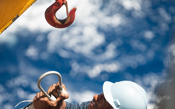 A construction or rigging worker wearing a white hard hat and blue work shirt is handling a metal shackle or lifting ring, apparently preparing to connect it to a crane hook visible in the upper left. The image is taken from a low angle looking upward against a partly cloudy blue sky. A second worker wearing a blue hard hat is partially visible in the lower right foreground.