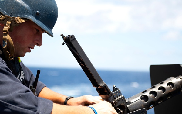 A U.S. Navy sailor in blue coveralls and a blue helmet is performing maintenance or assembly on a crew-served weapon system, likely an M2 .50 caliber machine gun, mounted on the deck of a naval vessel. The ocean is visible in the background under a partly cloudy sky, indicating an open-water operational environment. The sailor appears focused on the mechanical task at hand.