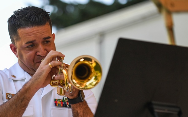 A U.S. Navy servicemember in white dress uniform is playing a brass trumpet while seated, reading from a music stand. He is wearing multiple service ribbons and rank insignia on his uniform, suggesting a formal or ceremonial performance. The setting appears to be an outdoor venue or covered stage.