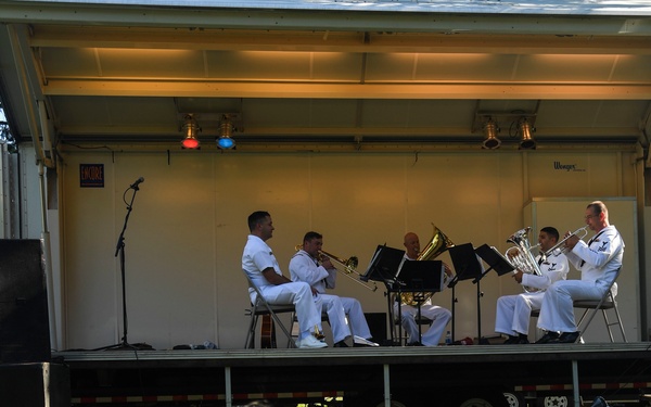 A military brass ensemble of approximately five musicians dressed in white naval uniforms performs on an outdoor mobile stage (trailer-mounted). Two audience members, an adult and a child, sit on the grass in the foreground watching the performance. The setting appears to be an outdoor public event during daylight hours.